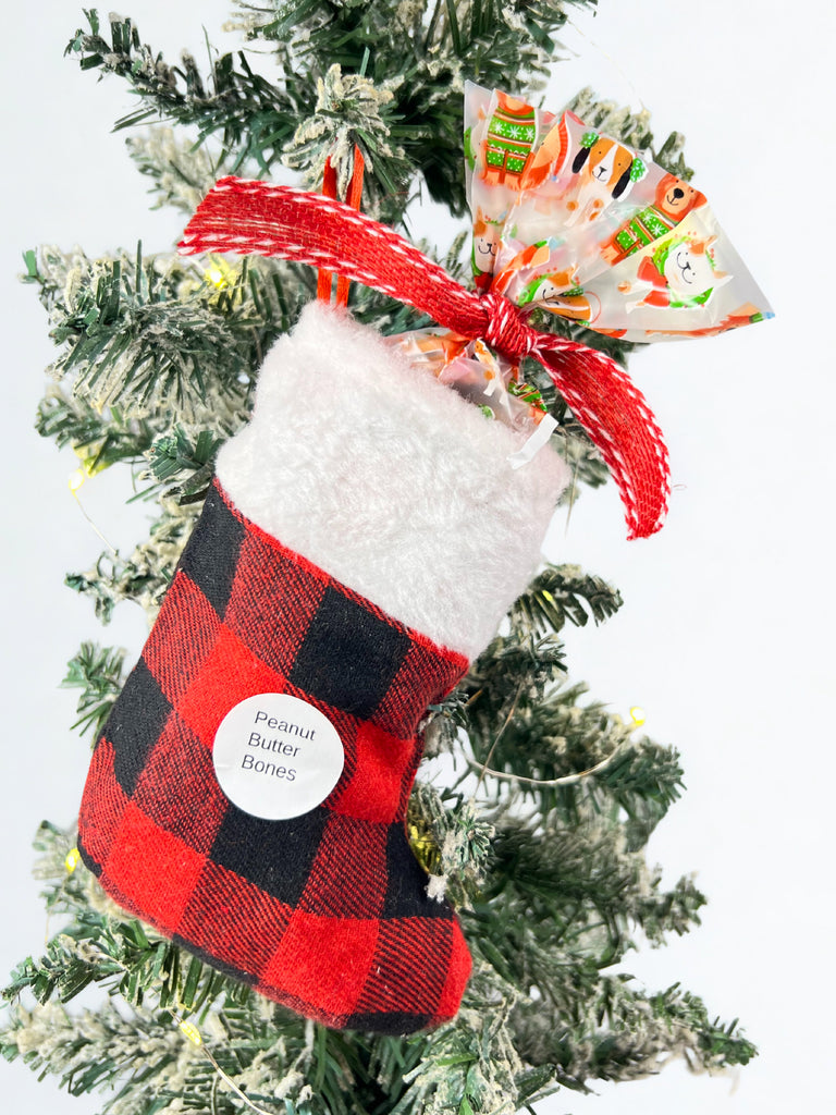 Red and black checkered stocking with a white fluffy interior, filled with wrapped gifts of dog peanut butter bones, hanging on a Christmas tree.