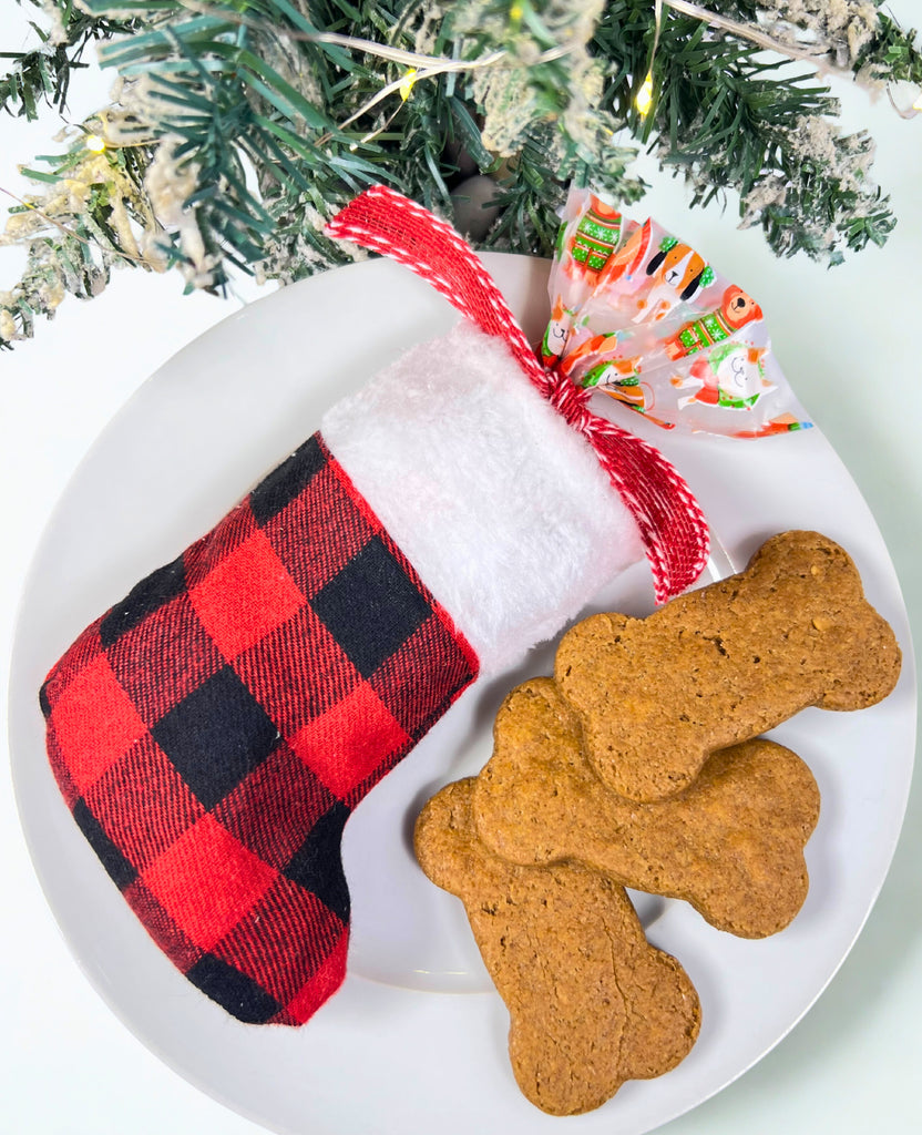 Dog treats shaped like bones on a white plate with a red and black checkered Christmas stocking and a Christmas tree in the background.
